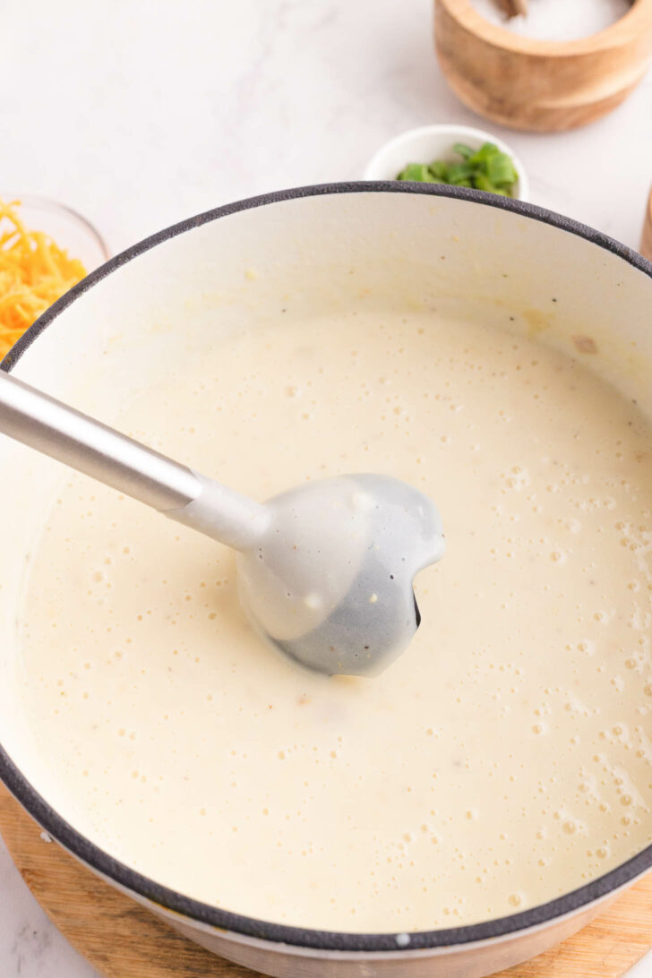 A large pot filled with creamy, light-colored soup, with an immersion blender partially submerged. Ingredients like shredded cheese and chopped green onions are visible in small bowls nearby.