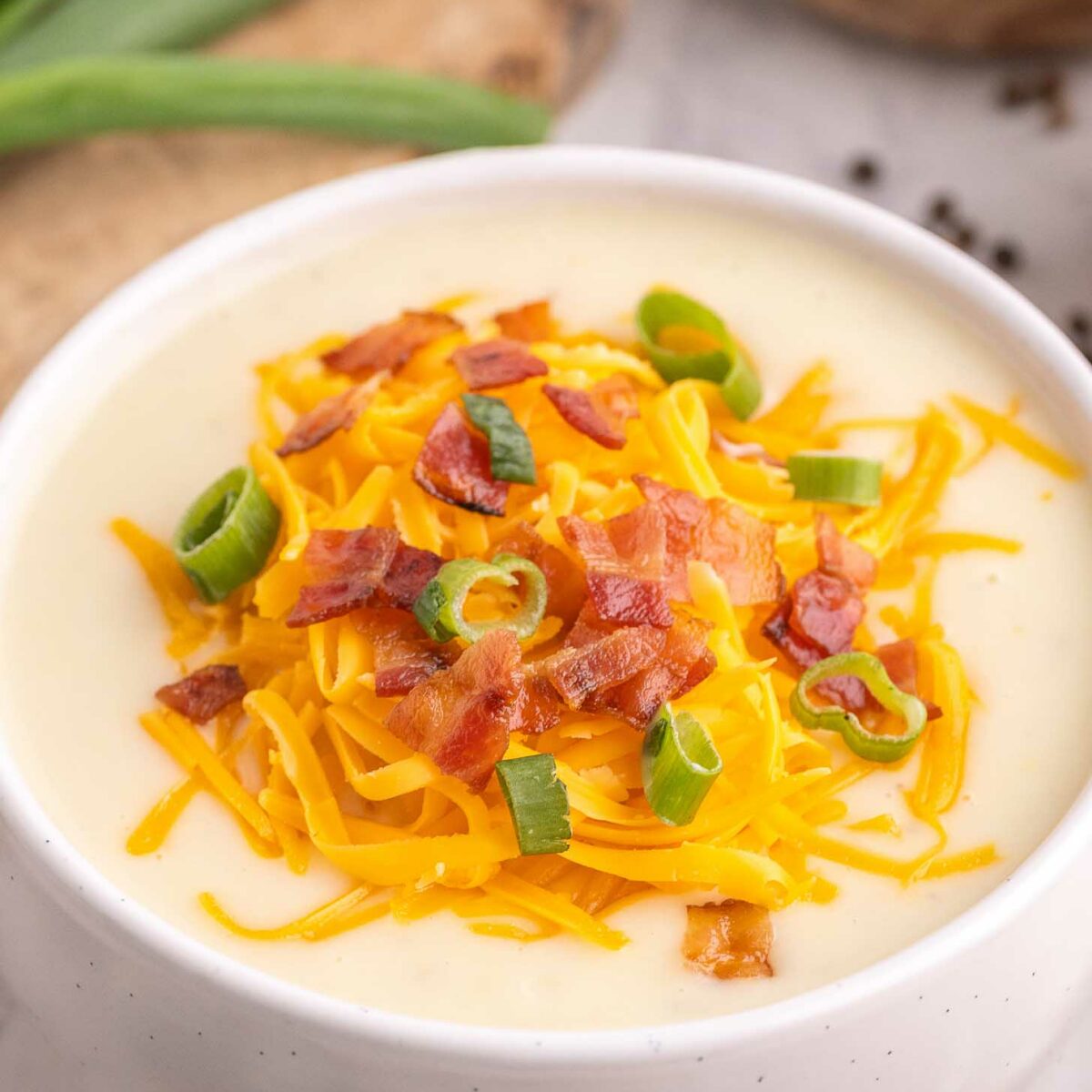 A white bowl filled with creamy soup topped with shredded cheddar cheese, crispy bacon pieces, and sliced green onions. Green onions and a wooden bowl are visible in the background.
