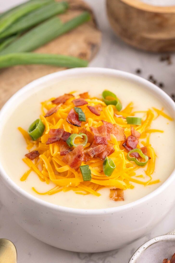 A white bowl filled with creamy soup topped with shredded cheddar cheese, crispy bacon pieces, and sliced green onions. Green onions and a wooden bowl are visible in the background.