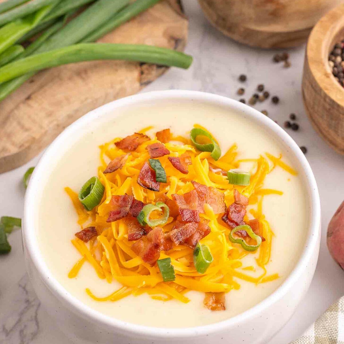A bowl of creamy potato soup topped with shredded cheddar cheese, crispy bacon bits, and sliced green onions. Sliced green onions, a bowl of salt, and a cutting board are in the background.