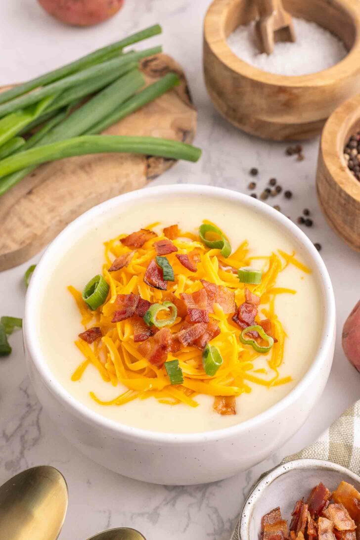 A bowl of creamy potato soup topped with shredded cheddar cheese, crispy bacon bits, and sliced green onions. Sliced green onions, a bowl of salt, and a cutting board are in the background.