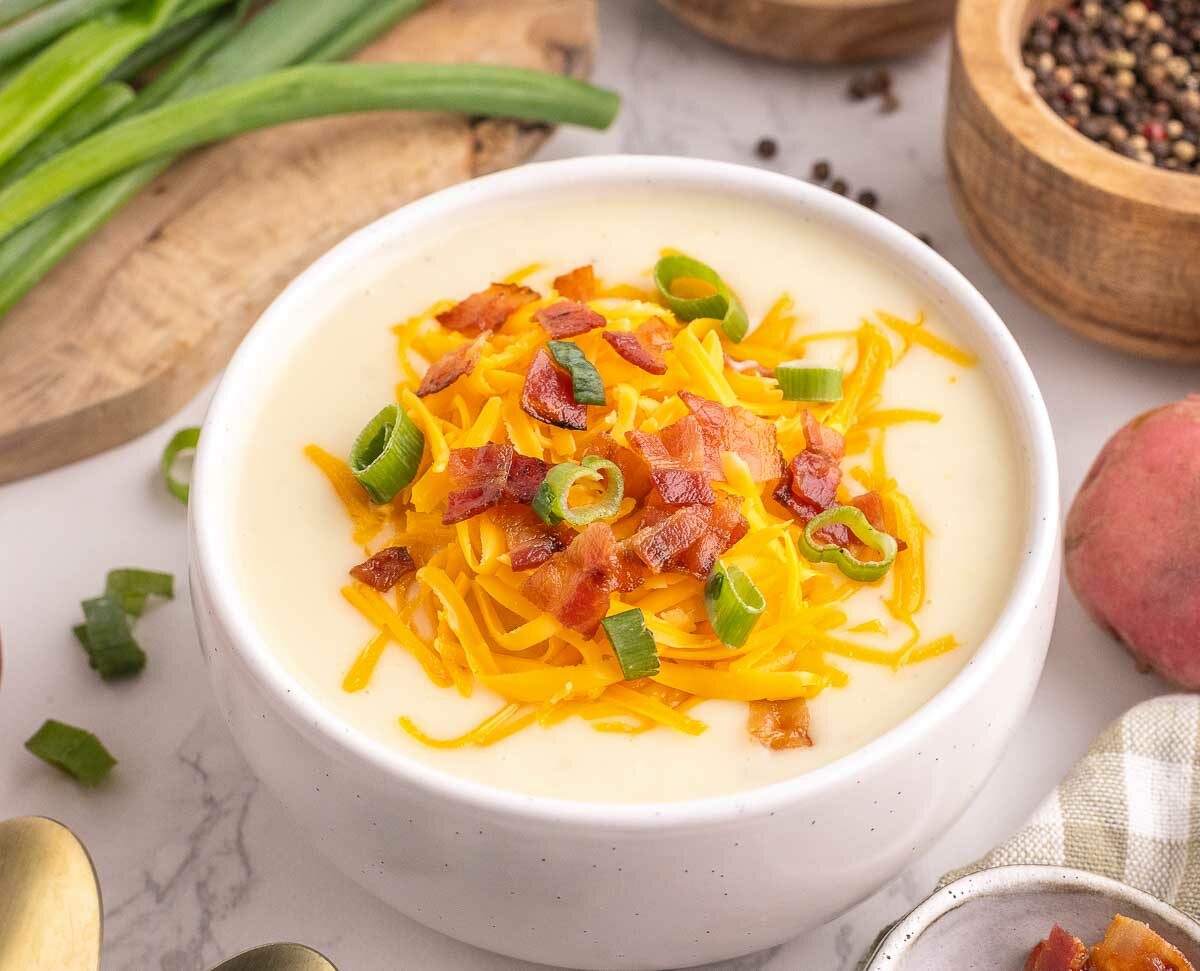 A bowl of creamy potato soup topped with shredded cheddar cheese, chopped bacon, and sliced green onions. Fresh potatoes, green onions, and bowls of spices are arranged around it on a white marble surface.