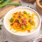 A bowl of creamy potato soup topped with shredded cheddar cheese, chopped bacon, and sliced green onions. Fresh potatoes, green onions, and bowls of spices are arranged around it on a white marble surface.