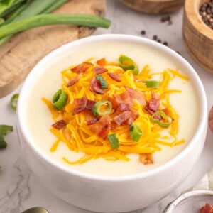 A bowl of creamy potato soup topped with shredded cheddar cheese, chopped bacon, and sliced green onions. Fresh potatoes, green onions, and bowls of spices are arranged around it on a white marble surface.