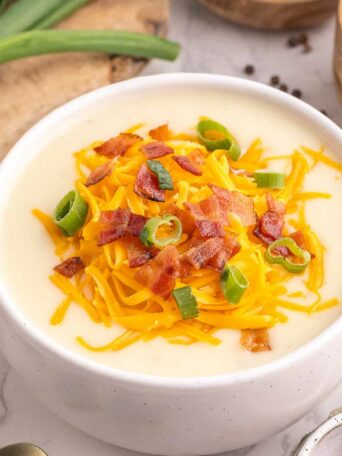 A bowl of creamy potato soup topped with shredded cheddar cheese, chopped bacon, and sliced green onions. Fresh potatoes, green onions, and bowls of spices are arranged around it on a white marble surface.