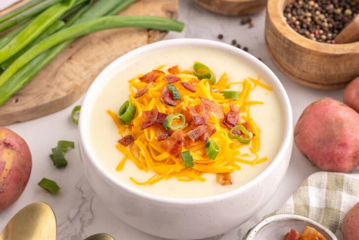 A bowl of creamy potato soup topped with shredded cheddar cheese, chopped bacon, and sliced green onions. Fresh potatoes, green onions, and bowls of spices are arranged around it on a white marble surface.