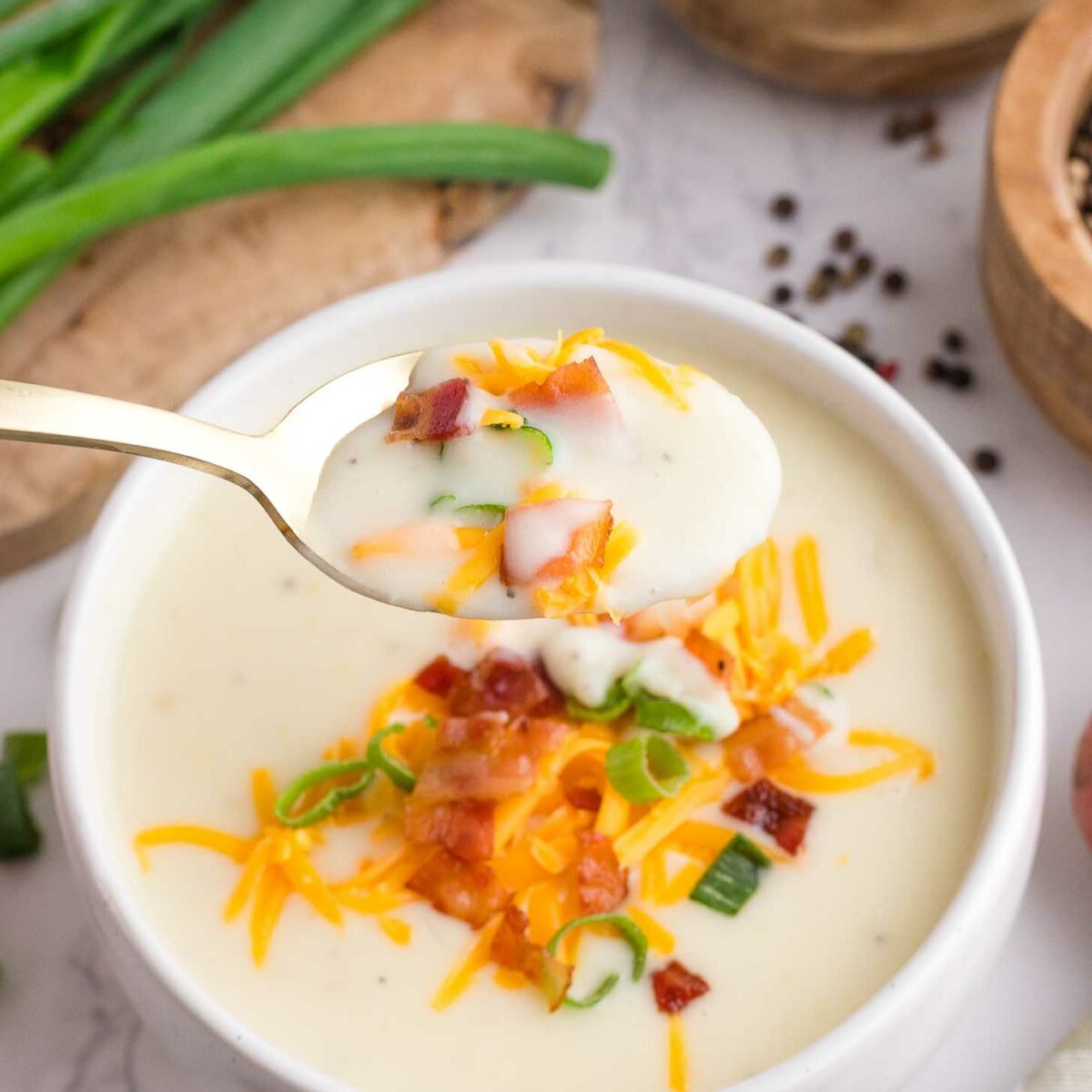 A bowl of creamy potato soup topped with shredded cheese, chopped green onions, and bacon pieces. A spoonful of the soup is held above the bowl, ready to eat. Green onions and seasonings are nearby.