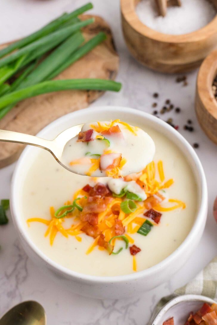 A bowl of creamy potato soup topped with shredded cheese, chopped green onions, and bacon pieces. A spoonful of the soup is held above the bowl, ready to eat. Green onions and seasonings are nearby.