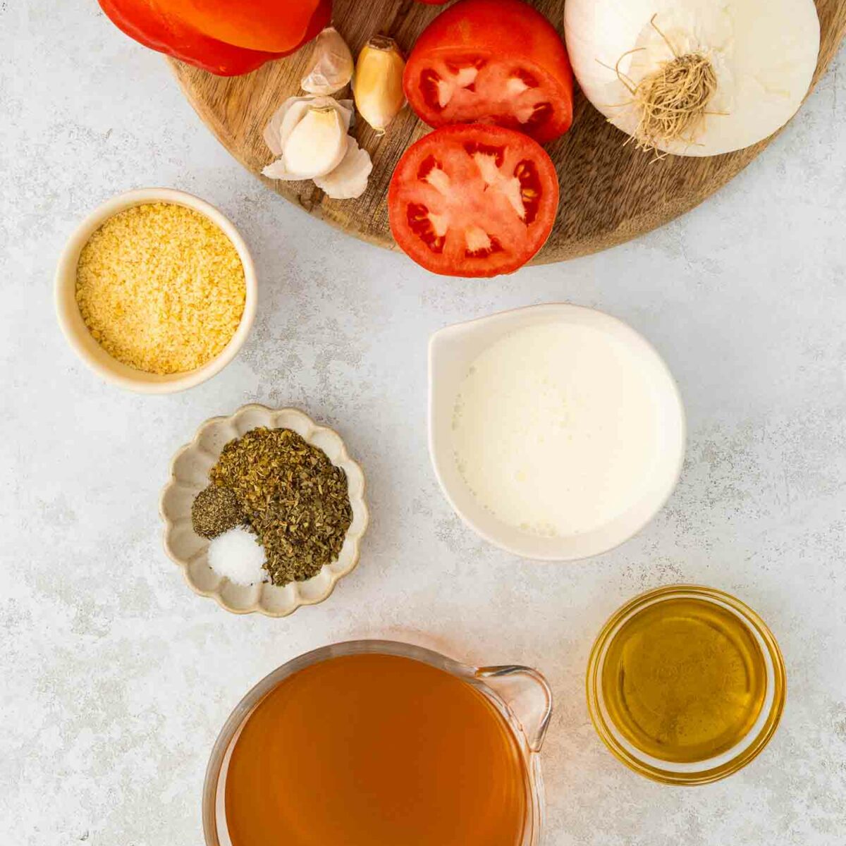 Ingredients for red pepper and tomato soup arranged on a white surface: fresh tomatoes, onion, garlic, grated cheese, dried herbs and salt, cream, olive oil, and a measuring cup of vegetable broth.