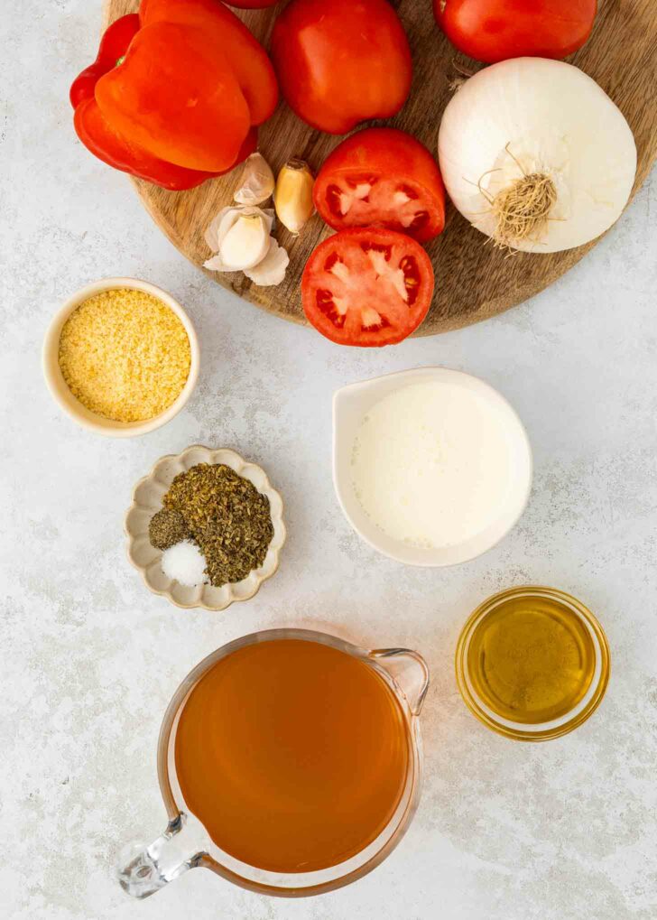 Ingredients for red pepper and tomato soup arranged on a white surface: fresh tomatoes, onion, garlic, grated cheese, dried herbs and salt, cream, olive oil, and a measuring cup of vegetable broth.