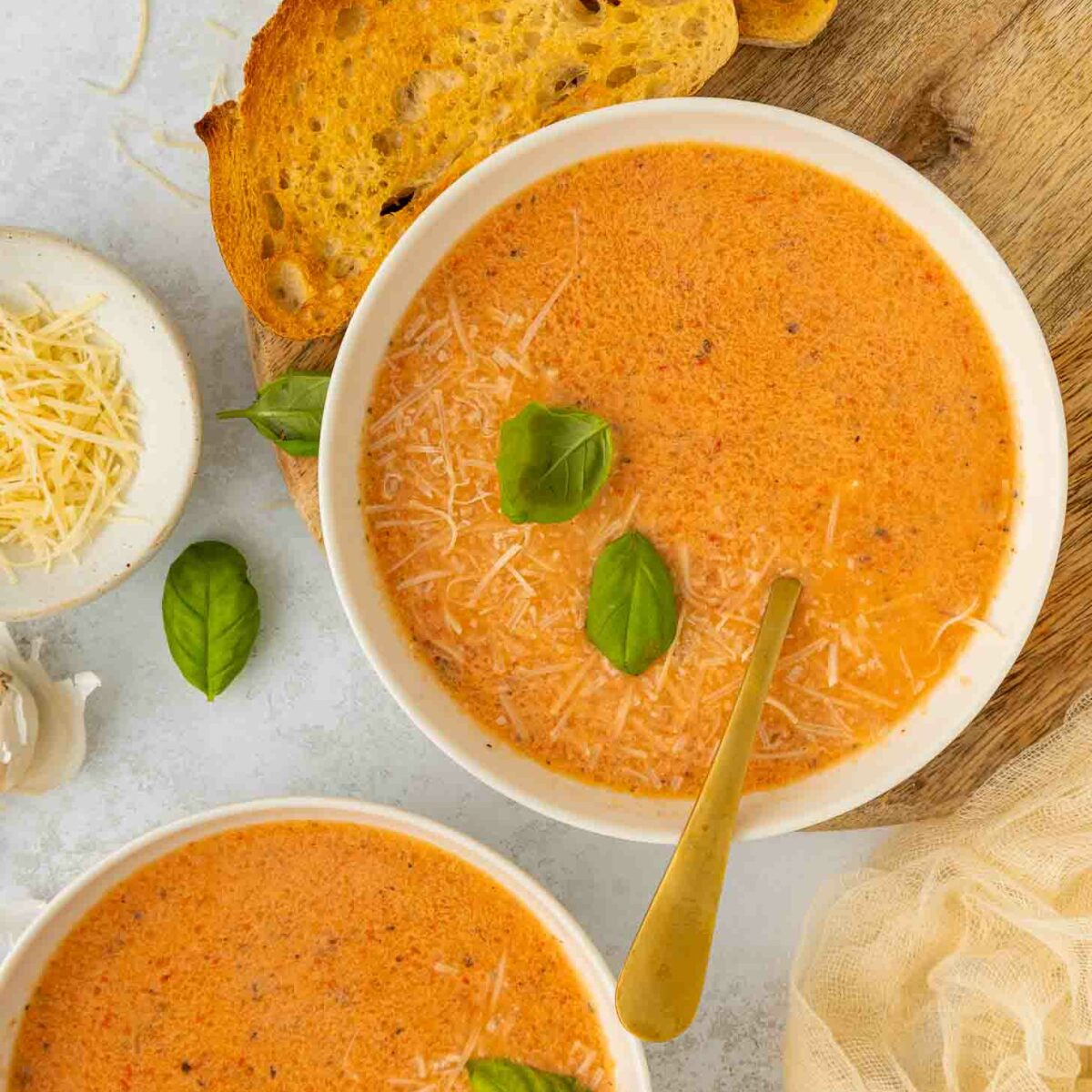 Two bowls of creamy red pepper and tomato soup topped with grated cheese and fresh basil leaves, served with slices of toasted bread on a wooden board. A small bowl of shredded cheese and a clove of garlic are nearby.