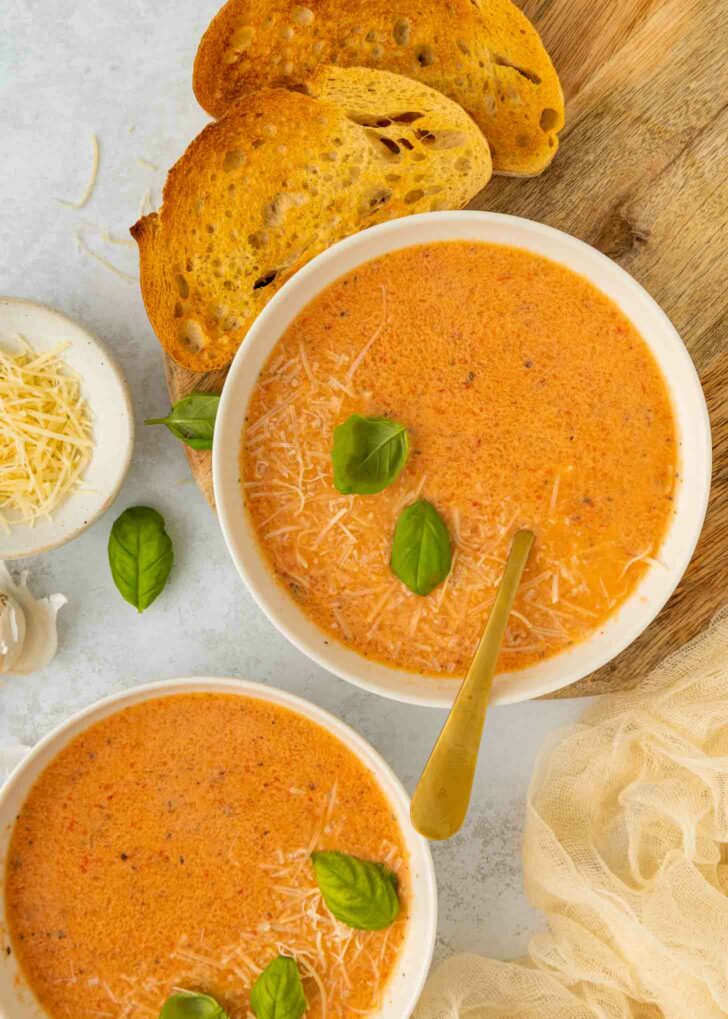 Two bowls of creamy red pepper and tomato soup topped with grated cheese and fresh basil leaves, served with slices of toasted bread on a wooden board. A small bowl of shredded cheese and a clove of garlic are nearby.