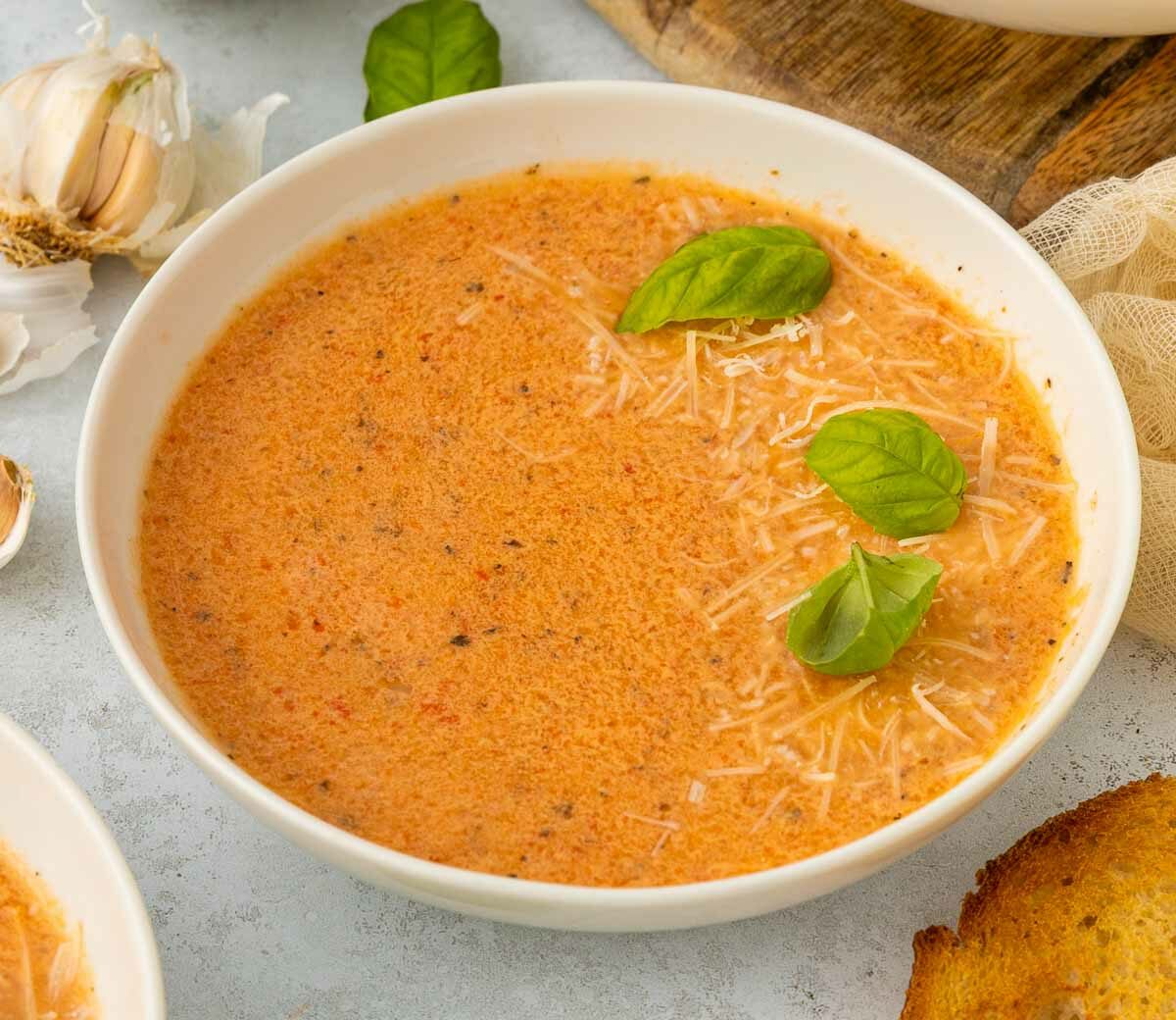 A bowl of creamy red pepper and tomato soup garnished with shredded cheese and fresh basil leaves, with garlic cloves and toasted bread nearby on a light surface.