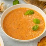 A bowl of creamy red pepper and tomato soup garnished with shredded cheese and fresh basil leaves, with garlic cloves and toasted bread nearby on a light surface.