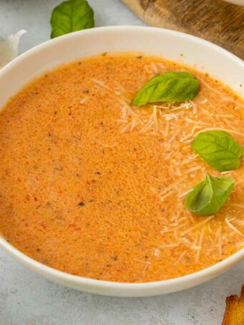 A bowl of creamy red pepper and tomato soup garnished with shredded cheese and fresh basil leaves, with garlic cloves and toasted bread nearby on a light surface.