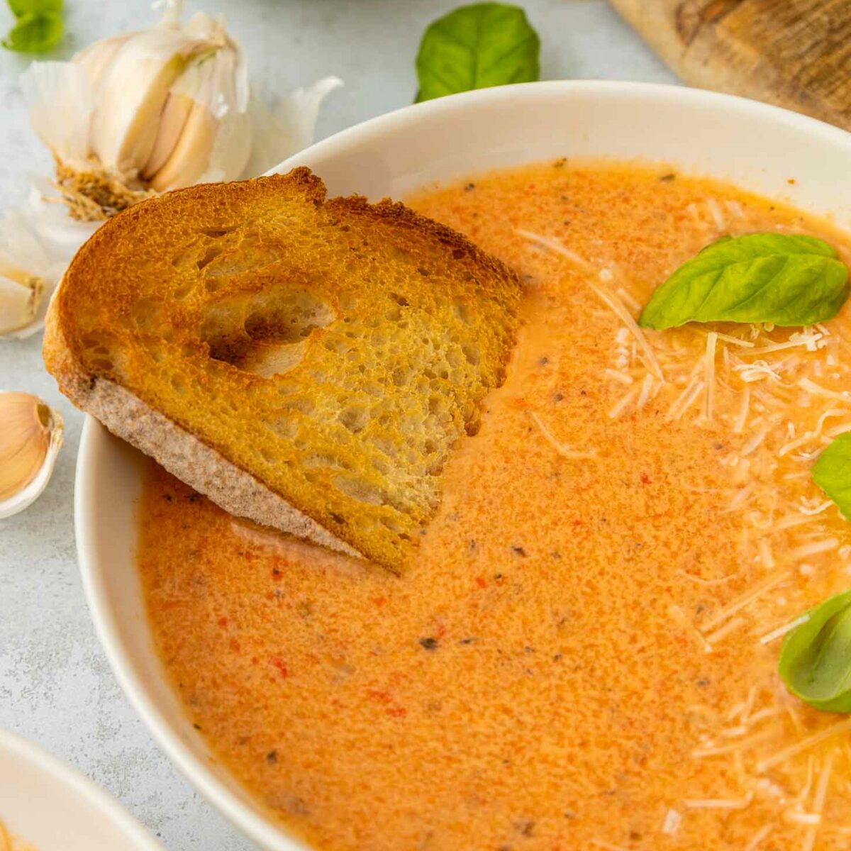 A bowl of creamy red pepper and tomato soup topped with shredded cheese and fresh basil leaves, served with a slice of toasted bread. Garlic bulbs and more cheese are visible in the background.