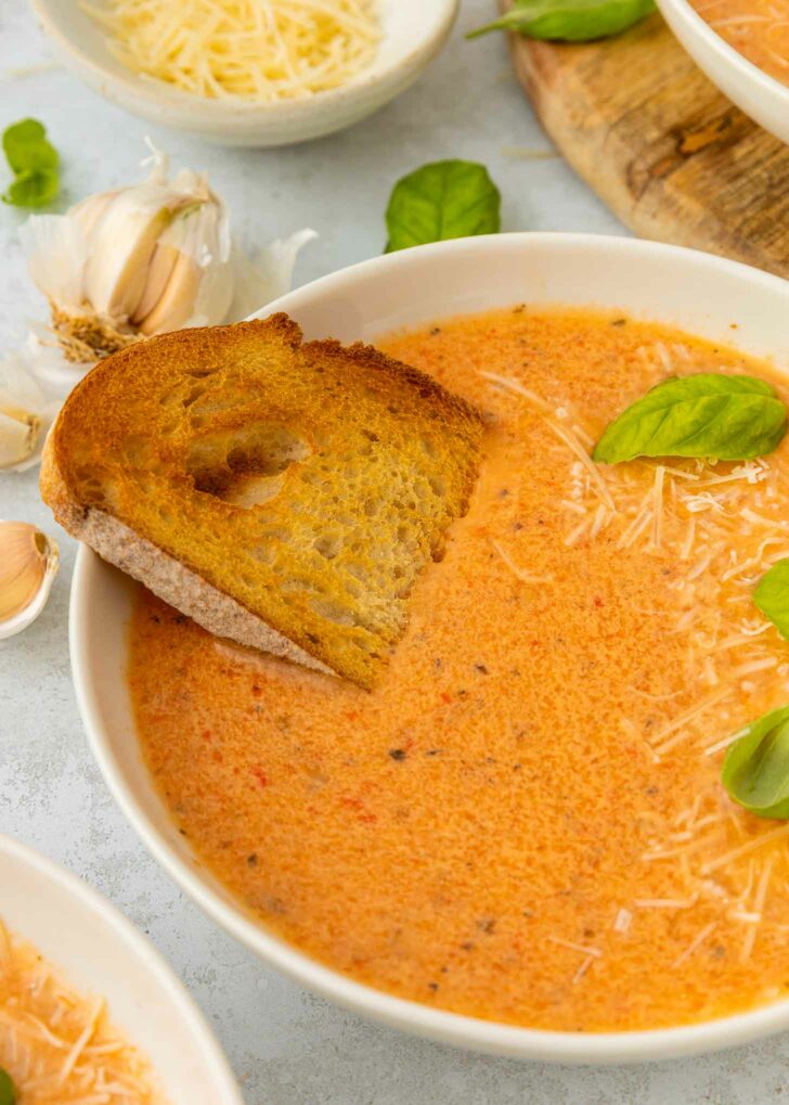 A bowl of creamy red pepper and tomato soup topped with shredded cheese and fresh basil leaves, served with a slice of toasted bread. Garlic bulbs and more cheese are visible in the background.