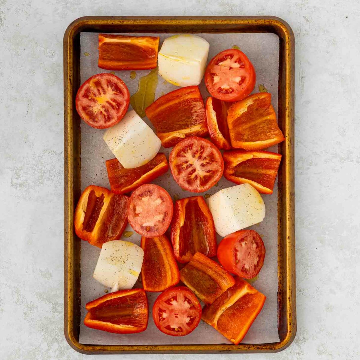 A baking tray with parchment paper holds chopped red bell peppers, halved tomatoes, and large chunks of onion, all drizzled with oil and sprinkled with pepper, ready to be roasted.