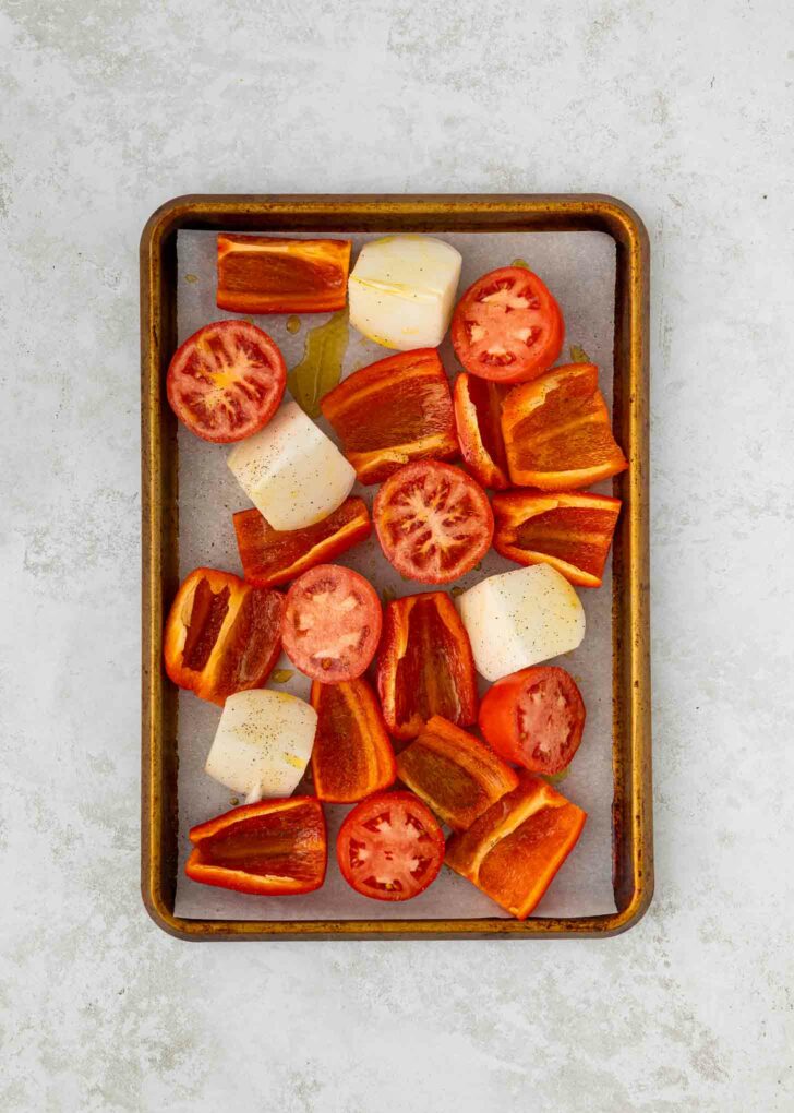 A baking tray with parchment paper holds chopped red bell peppers, halved tomatoes, and large chunks of onion, all drizzled with oil and sprinkled with pepper, ready to be roasted.