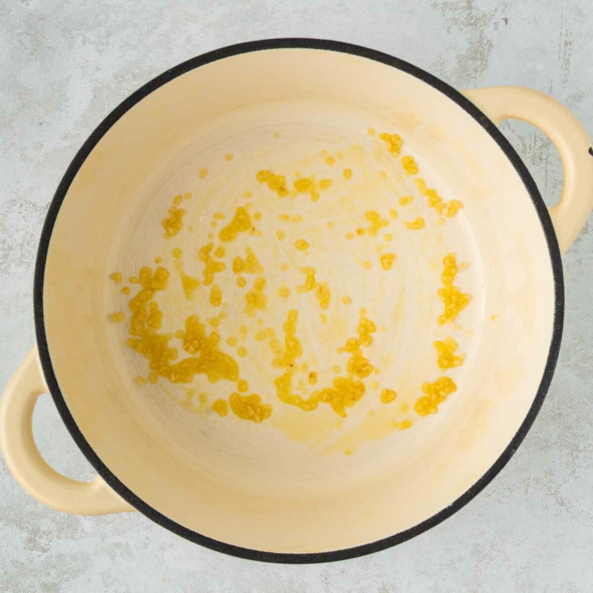 A large cream-colored pot with two handles contains a small amount of oil and garlic spread across the bottom. The pot is placed on a light gray textured surface.