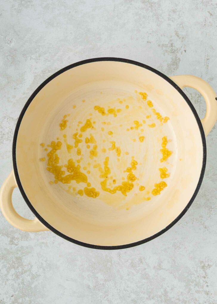 A large cream-colored pot with two handles contains a small amount of oil and garlic spread across the bottom. The pot is placed on a light gray textured surface.