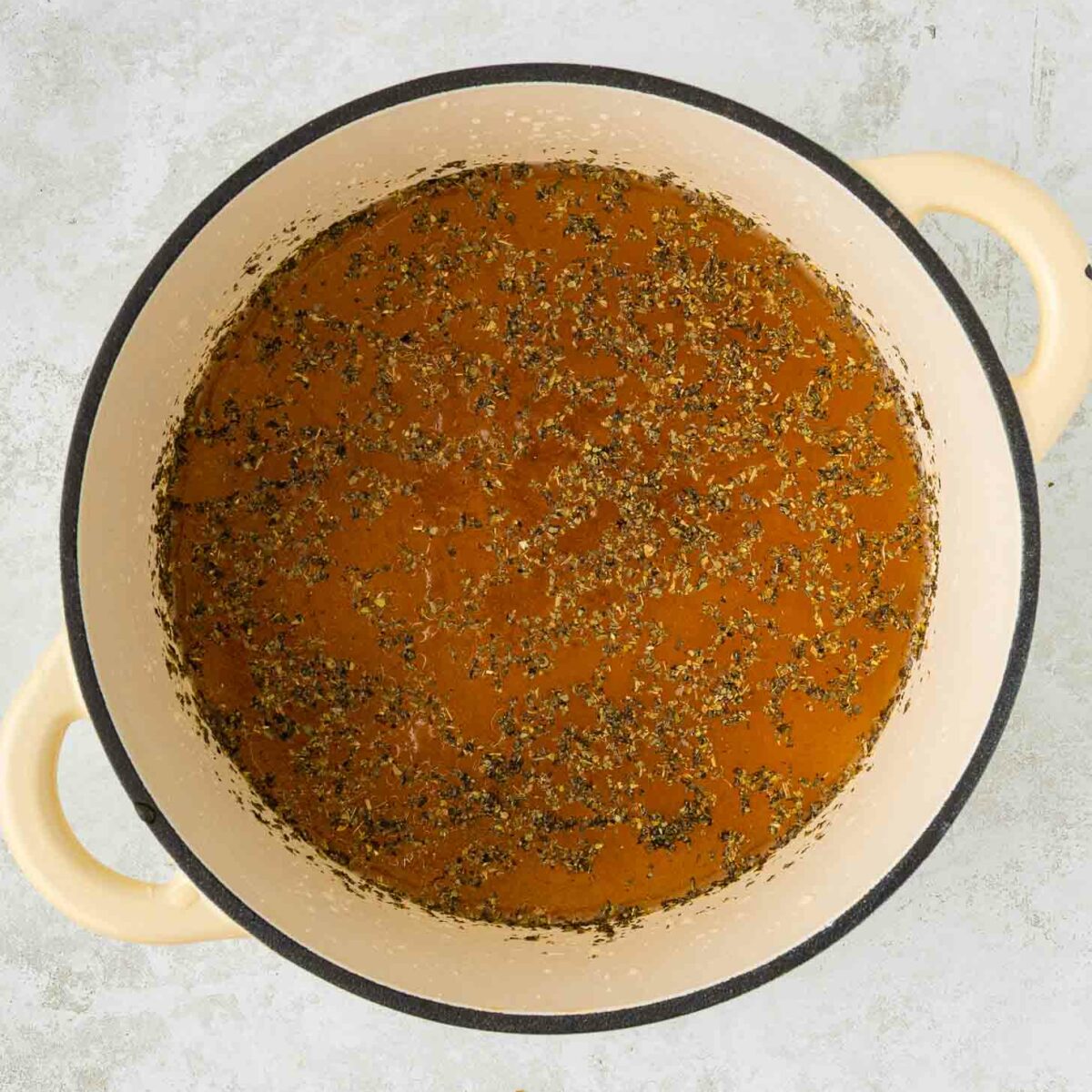 A beige Dutch oven filled with seasoned brown broth sits on a light gray textured surface.
