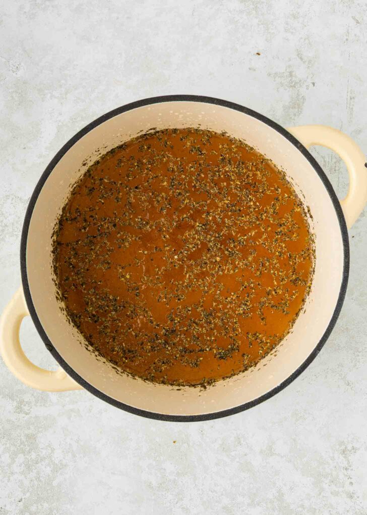 A beige Dutch oven filled with seasoned brown broth sits on a light gray textured surface.