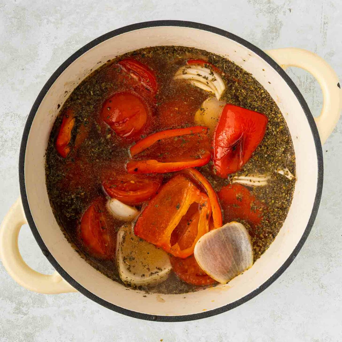 A cream-colored pot filled with tomato halves, red bell pepper pieces, garlic cloves, herbs, and broth, all simmering together on a light gray background.