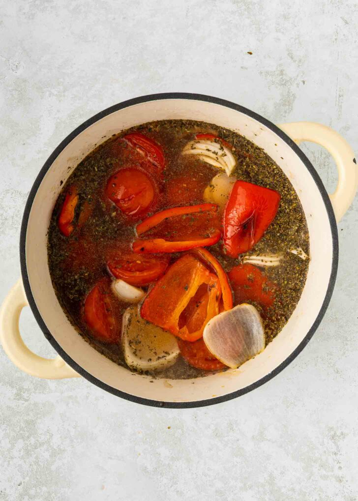 A cream-colored pot filled with tomato halves, red bell pepper pieces, garlic cloves, herbs, and broth, all simmering together on a light gray background.