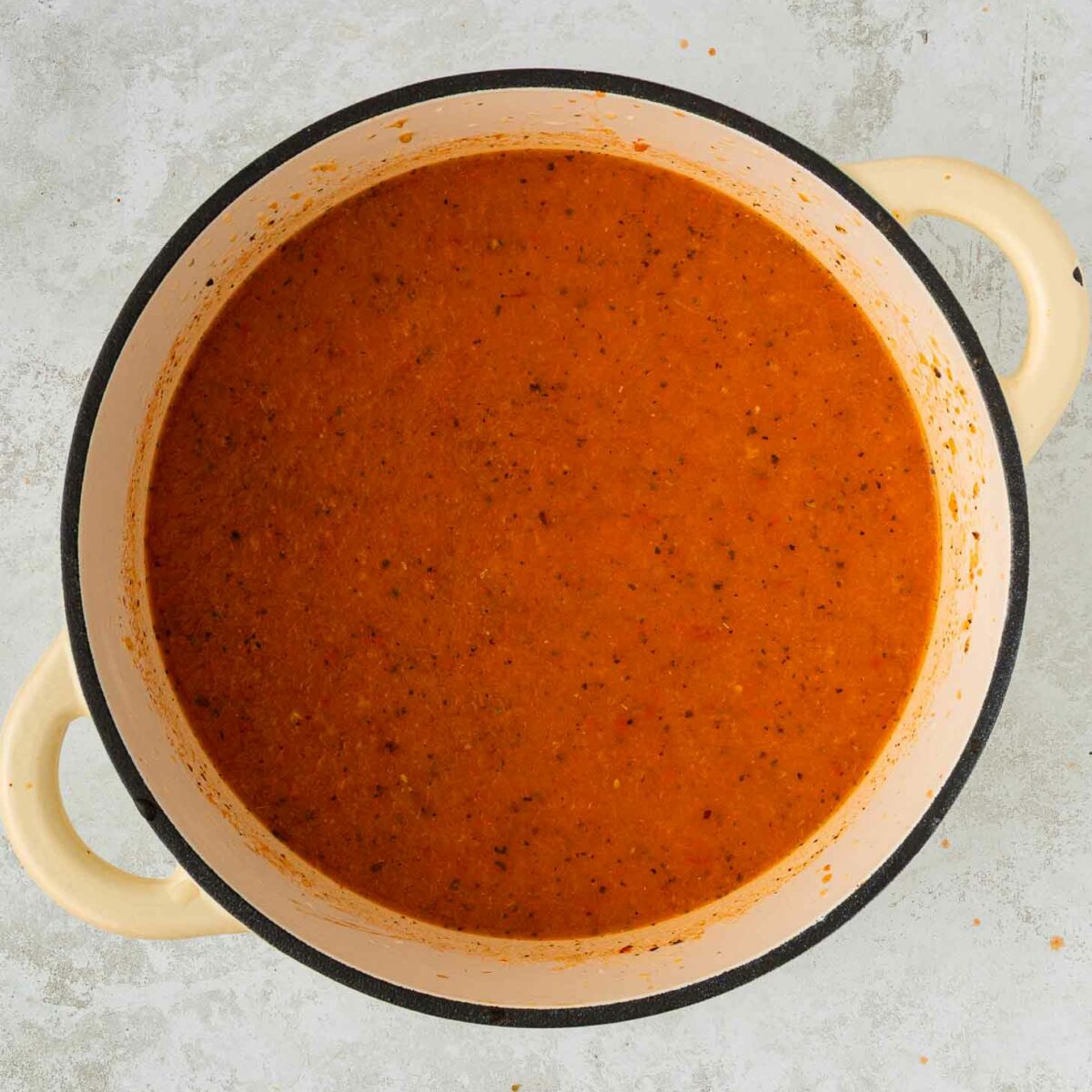 A cream-colored pot filled with smooth, orange-red tomato soup, viewed from above on a light gray textured surface.