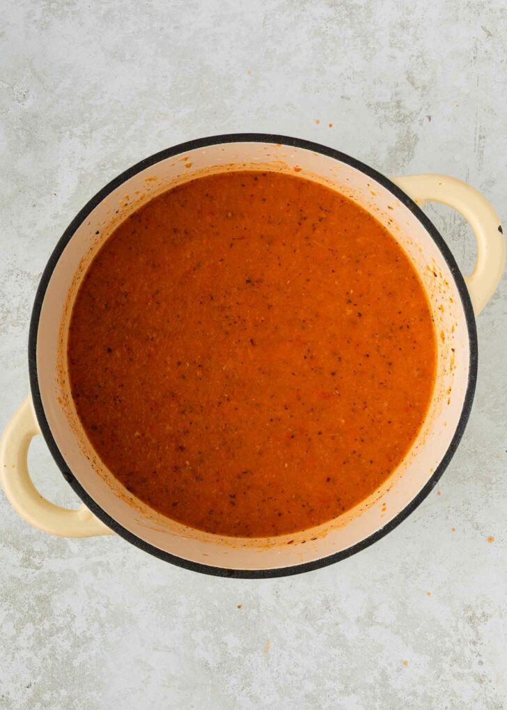 A cream-colored pot filled with smooth, orange-red tomato soup, viewed from above on a light gray textured surface.