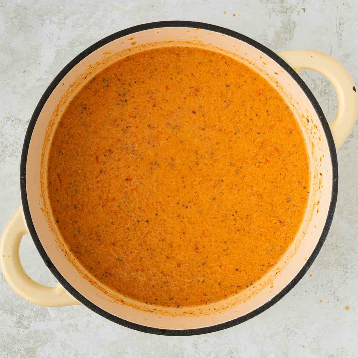 A large cream-colored pot filled with creamy, orange-colored soup or sauce, speckled with herbs and spices, sits on a light gray surface.