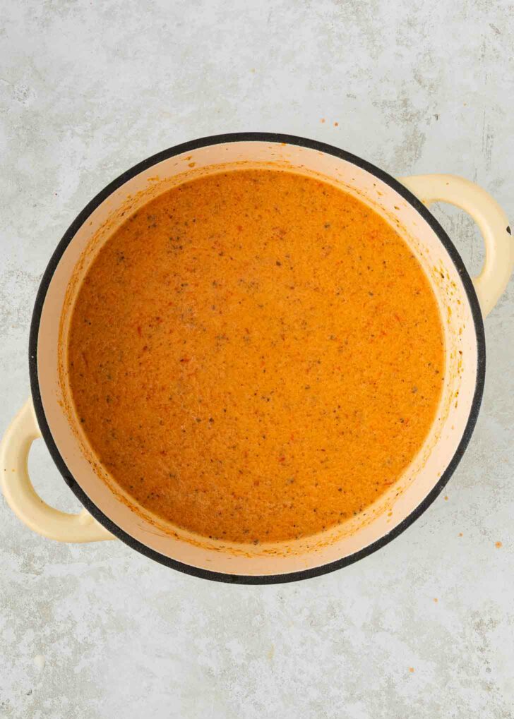 A large cream-colored pot filled with creamy, orange-colored soup or sauce, speckled with herbs and spices, sits on a light gray surface.
