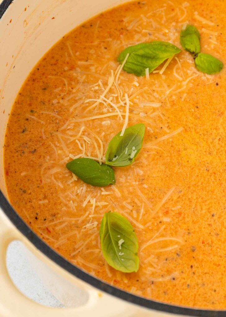 A creamy orange soup garnished with fresh basil leaves and shredded Parmesan cheese, served in a white pot. The soup surface shows flecks of herbs and a smooth, rich texture.