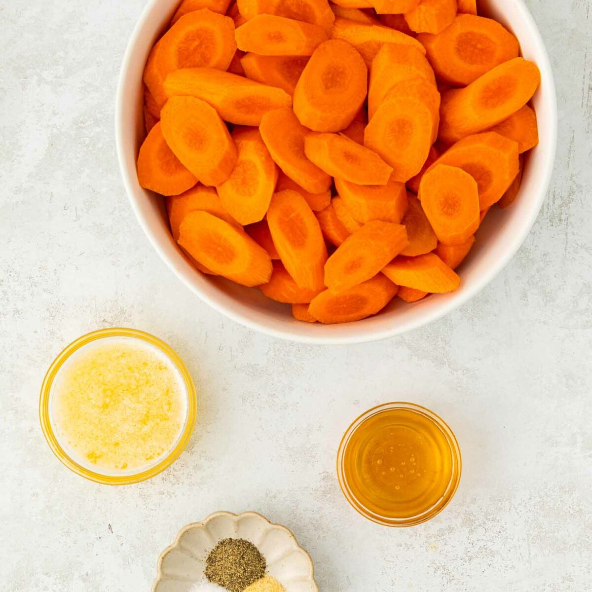 A bowl of sliced carrots is surrounded by small bowls containing melted butter, honey, and a dish with salt, pepper, and garlic powder on a light surface.