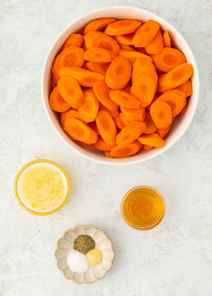 A bowl of sliced carrots is surrounded by small bowls containing melted butter, honey, and a dish with salt, pepper, and garlic powder on a light surface.