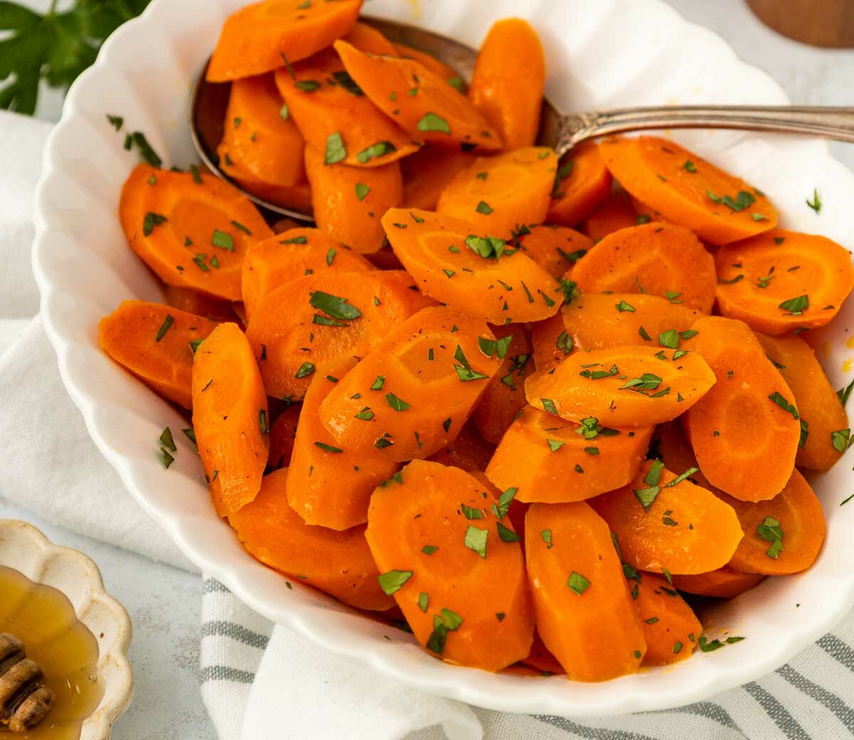 A white bowl filled with sliced cooked carrots garnished with chopped herbs, with a serving spoon on the side. A small dish of honey and a white cloth are visible in the background.