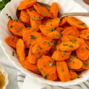 A white bowl filled with sliced cooked carrots garnished with chopped herbs, with a serving spoon on the side. A small dish of honey and a white cloth are visible in the background.