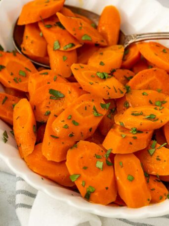A white bowl filled with sliced cooked carrots garnished with chopped herbs, with a serving spoon on the side. A small dish of honey and a white cloth are visible in the background.