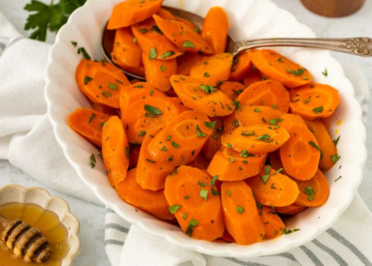 A white bowl filled with sliced cooked carrots garnished with chopped herbs, with a serving spoon on the side. A small dish of honey and a white cloth are visible in the background.