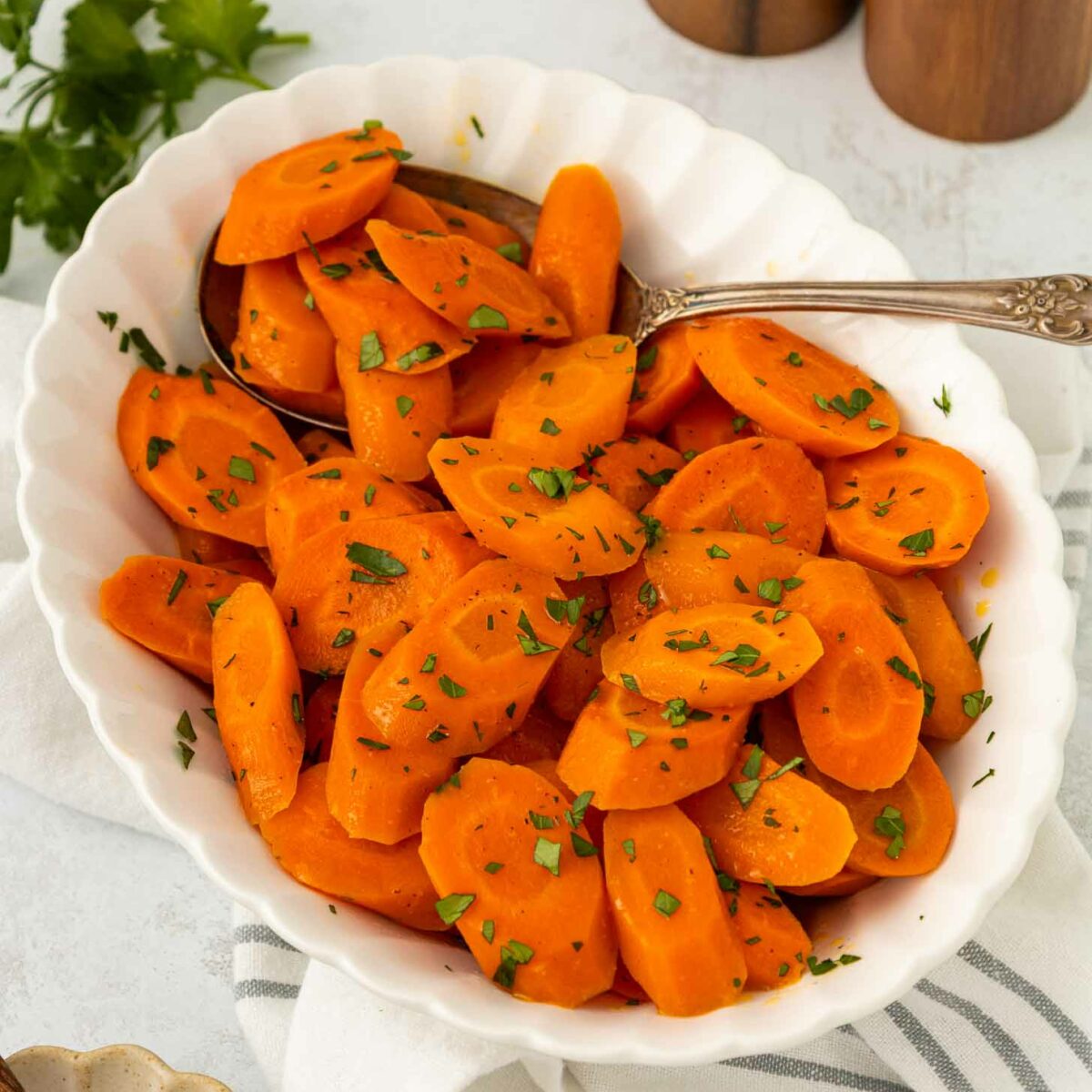 A white bowl filled with sliced cooked carrots garnished with chopped parsley, with a spoon resting in the bowl. Fresh parsley, a striped napkin, and a honey dipper are nearby.