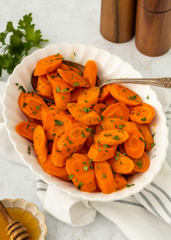 A white bowl filled with sliced cooked carrots garnished with chopped parsley, with a spoon resting in the bowl. Fresh parsley, a striped napkin, and a honey dipper are nearby.