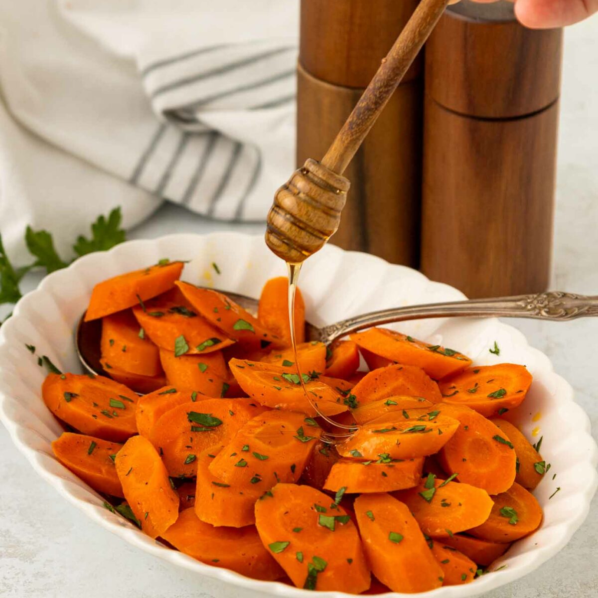 A hand drizzles honey over a bowl of sliced cooked carrots garnished with chopped herbs. A spoon rests in the dish, with salt and pepper shakers and a striped towel in the background.