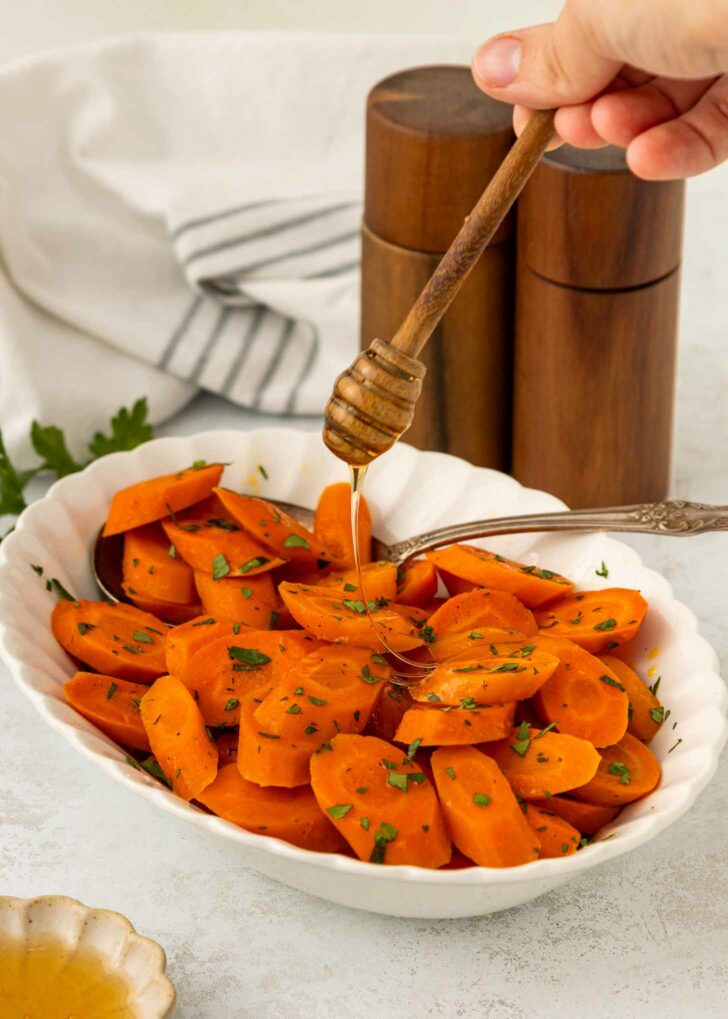 A hand drizzles honey over a bowl of sliced cooked carrots garnished with chopped herbs. A spoon rests in the dish, with salt and pepper shakers and a striped towel in the background.