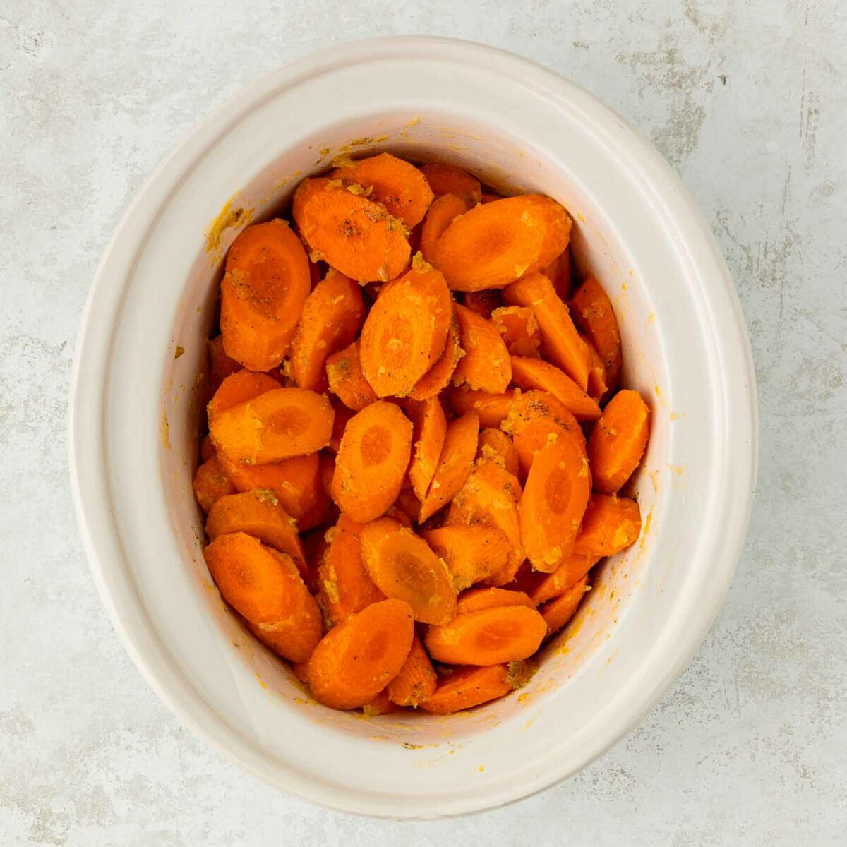 A white ceramic bowl filled with sliced cooked carrots, seasoned and arranged in a small pile, sits on a light textured surface.