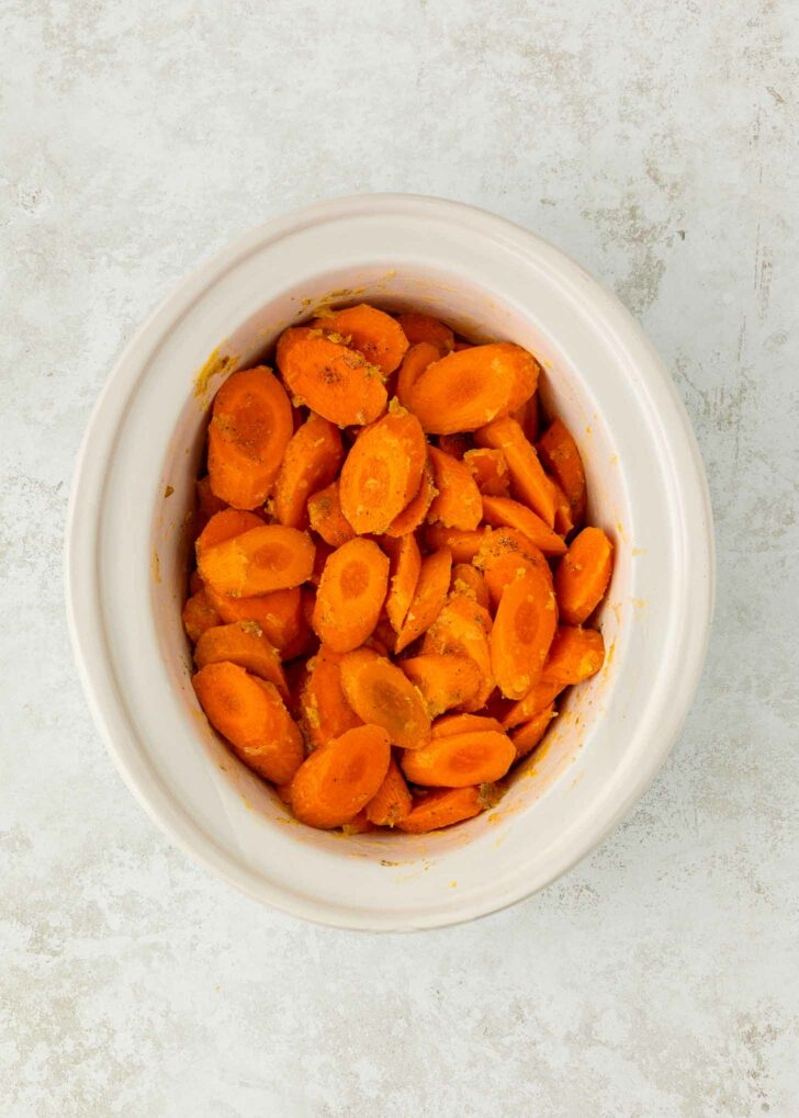 A white ceramic bowl filled with sliced cooked carrots, seasoned and arranged in a small pile, sits on a light textured surface.
