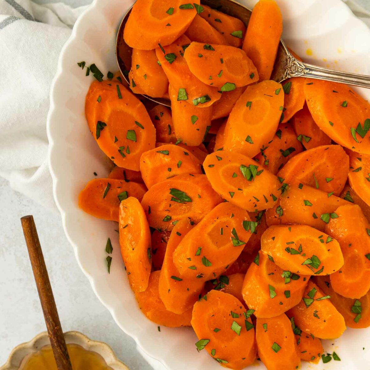 A white bowl filled with sliced cooked carrots garnished with chopped herbs. A silver serving spoon rests in the bowl. Nearby, there is a dish of honey with a honey dipper.