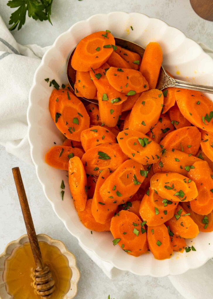 A white bowl filled with sliced cooked carrots garnished with chopped herbs. A silver serving spoon rests in the bowl. Nearby, there is a dish of honey with a honey dipper.