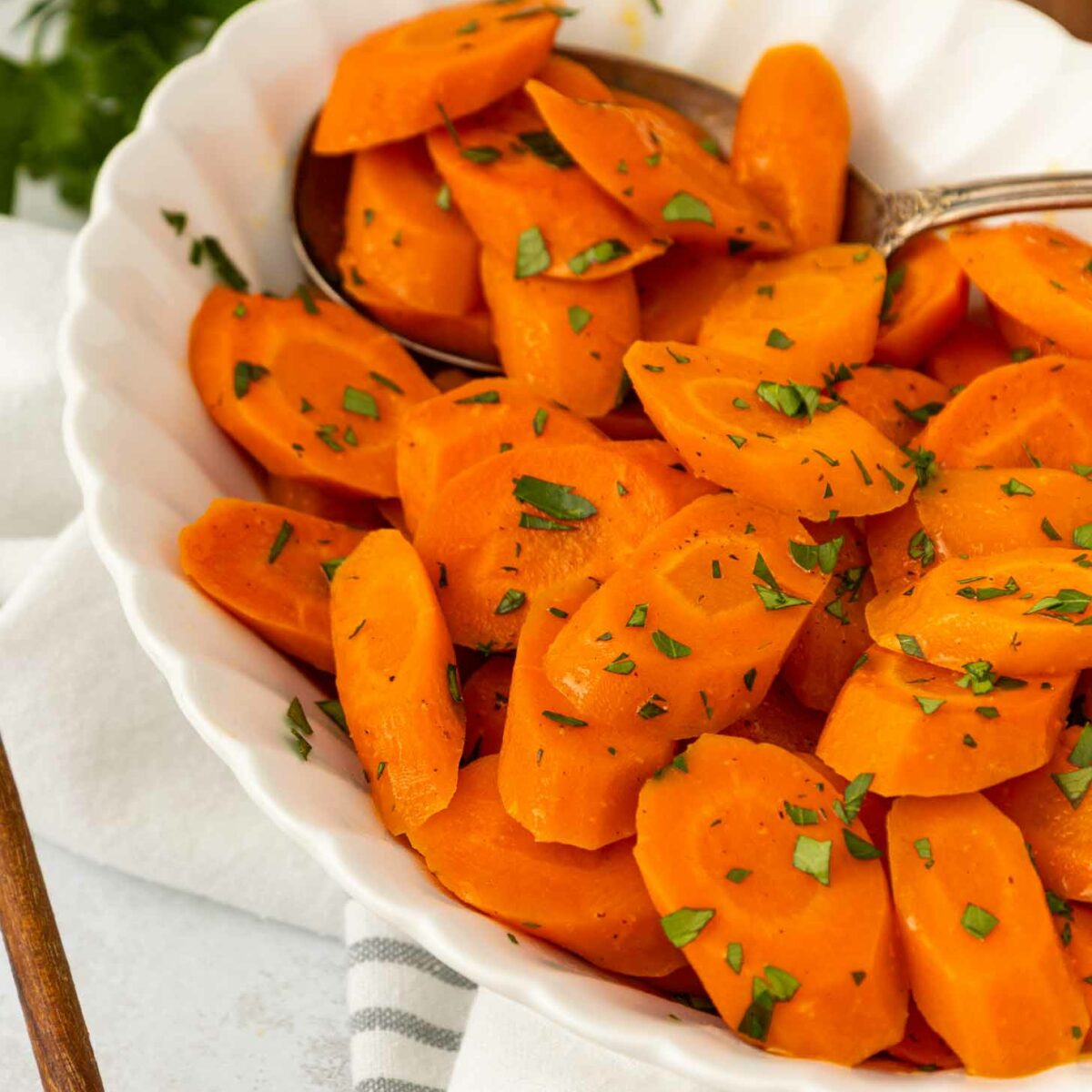 A white bowl filled with sliced cooked carrots, garnished with chopped parsley, with a spoon inside. The bowl rests on a striped cloth with a leafy garnish in the background.