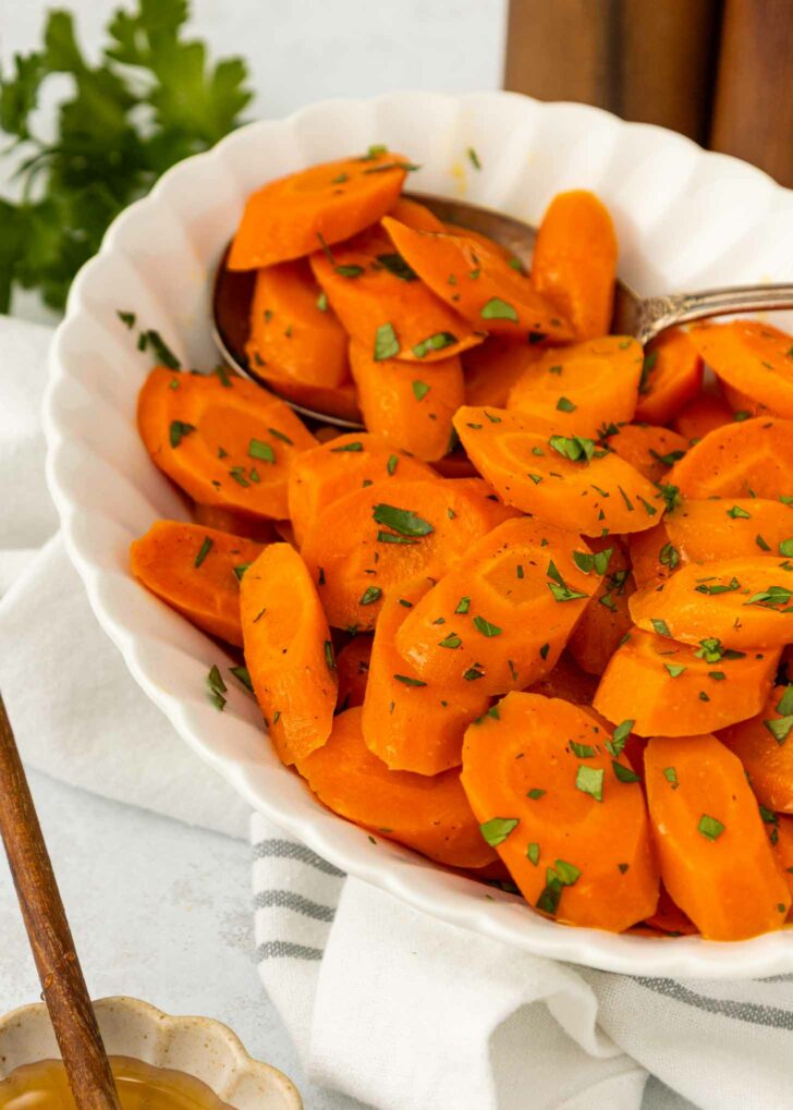 A white bowl filled with sliced cooked carrots, garnished with chopped parsley, with a spoon inside. The bowl rests on a striped cloth with a leafy garnish in the background.